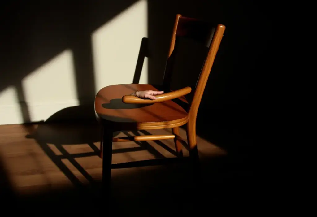 Une chaise en bois dans une pièce sombre, mains posées sur le bras