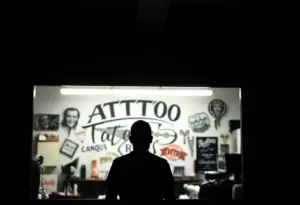 Une silhouette observe son reflet dans un miroir d'un studio de tatouage, entouré d'œuvres et d'outils