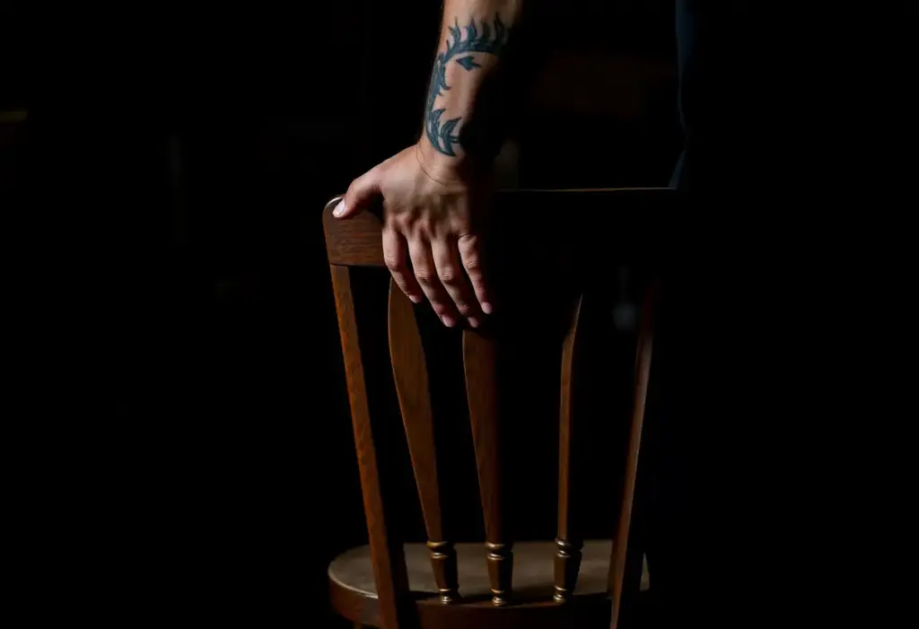 Main de l'homme tenant une chaise en bois dans une pièce sombre, avant un tatouage